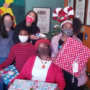 Children holding presents and an AEG employee surround a sitting mother, all wearing masks and smiling. 