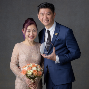 Two people stood next to each other smiling, one person holding flowers and the other holding an award