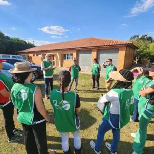 Students and adults in matching green vests meet in a circle outside a building in a rural area.
