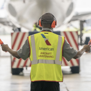 A person guiding a plane wearing a hi-vis vest that reads "American aircraft maintenance" 