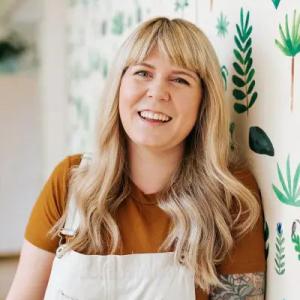 Ally Watson, smiling, leaning on a wall with painted plants.