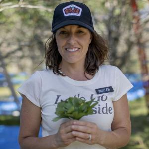 Alyssa in a white t-shirt and dark baseball cap holding a small bunch of greenery