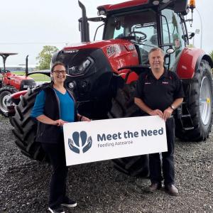 Two people stand in front of a tractor with a "Meet the Need" sign