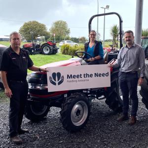 Three people stand with front of a tractor with a "Meet the Need" sign