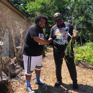 Two people shaking hands while looking at the camera. One holds a bundle of produce. They're standing on a mulch path in a vegetable garden.