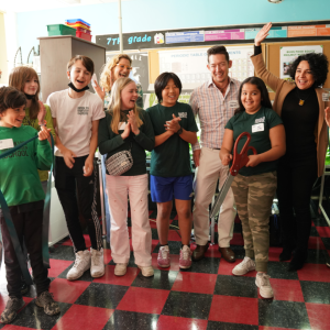 A group of adults and children are standing in a classroom clapping as a student cuts ribbon with oversized scissors. 