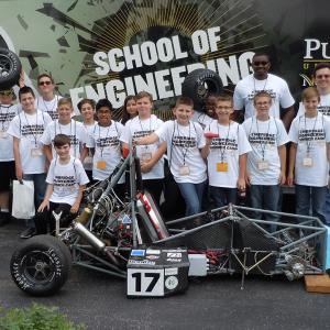 Large group of students and adults stand behind a go-cart type vehicle. All wearing the same 'school of engineering' t shirt