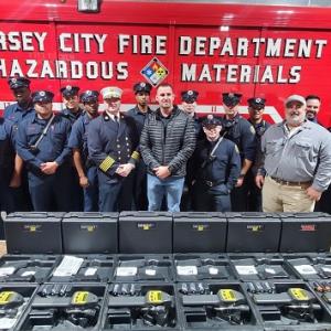 A group of 16 people in uniform stand in front of a Large red vehicle "Jersey City Fire Department Hazardous Materials" on the side. They stand behind 12 cases of equipment.