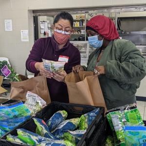two people look at a bag of vegetables, one holds an open paper bag. They are in a kitchen type setting with crates of bagged vegetables in front of them