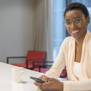 person sitting at a large table with phone in both hands and coffee mug next to them, smiling at the camera