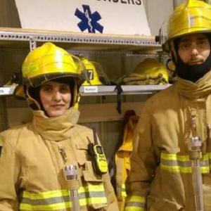 Two people in firefighter uniforms stand in front of a closet of safety equipment