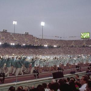 athletes dressed in matching uniforms walking along a track of a stadium. Stands filled with spectators.