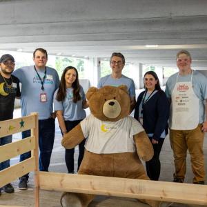 A group of volunteers posed with a large teddy bear and a bed frame.
