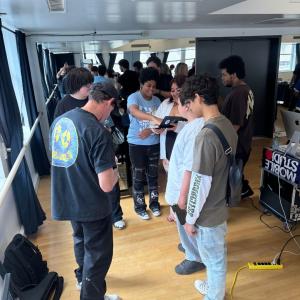 A group gathers around music and production equipment during a hands-on studio program, with additional participants and equipment visible throughout the room.