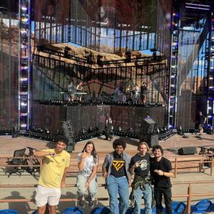 Five people pose in front of the stage at Red Rocks Amphitheatre while production crew and equipment are visible onstage behind them.