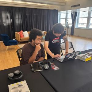 Two people work together at a laptop during a music program, with AXS-branded headphones, Skullcandy products and program materials displayed on a table.