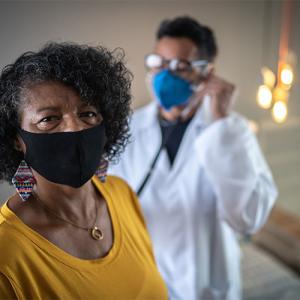 A medical professional using a stethoscope on a patient who is looking at the camera. Both are wearing protective masks.