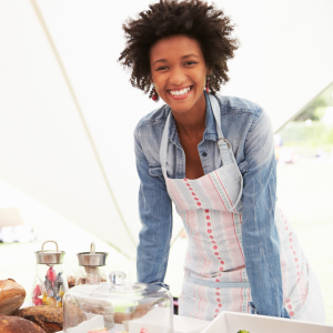 a person wearing an apron in a white tent, baked goods in front of them