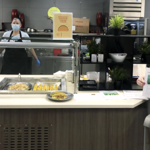 2 Blessing Hospital System masked employees stand behind a taco serving area in a cafeteria. An additional employee sits in front of the station, masked, holding up a document