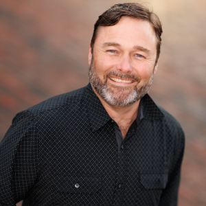 Man with a beard and mustache wearing a dark patterned button-up shirt, smiling in front of a blurred brown and gray background.