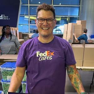Brock Carlson, Manager- Talent Acquisition FedEx Logistics, stood in front of boxes wearing a purple fedEx cares photo
