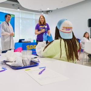 Students sit at a big table with paper and pencils while they talk with scientists at the head of a classroom.