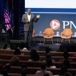 Richard Bynum standing on a stage in an auditorium of seated people. Chairs and PNC banner behind them.