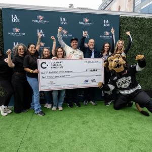 Group stands on a branded step-and-repeat holding a large donation check for the Culinary Careers Program at LA LIVE, with a mascot posing nearby.