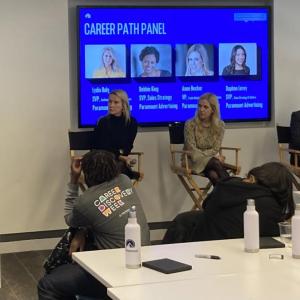 Four people seated with a display "Career path panel" behind them, all listening to a speaker at a podium to the left. Students seated at tables in the room.