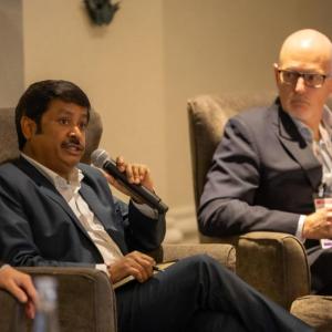 Two men in business attire are seated in armchairs during a conference. One holds a microphone and is speaking, while the other looks on attentively. Both wear name badges and are engaged in a discussion