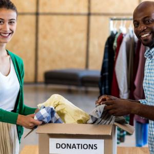 Two people holding clothing items in a "Donations" box. A rack of clothing hanging behind them.