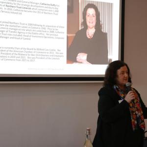 Catherine speaking to the room, a large display behind them.