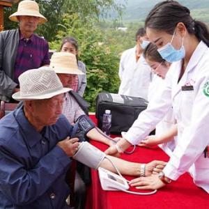 Two elderly people sit at a table outside and have their blood pressure taken by two people in lab coats. A queue of others behind them.