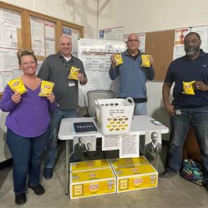 Four people holding up bags of chips at a small table.