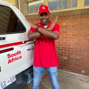Man in a red shirt next to a van that says South Africa, crossing his arms
