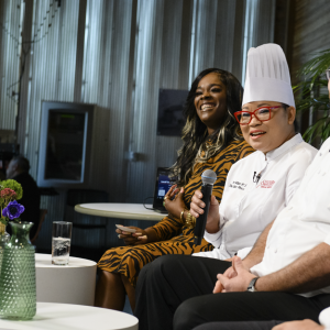 2 male chefs look at a female chef speaking as a part of a panel. The panel facilitator looks out into the audience.