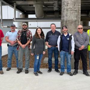 Eight people stand in a row outside . Large cement pillars behind them