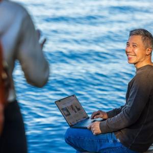 Person on a laptop seated next to a lake.