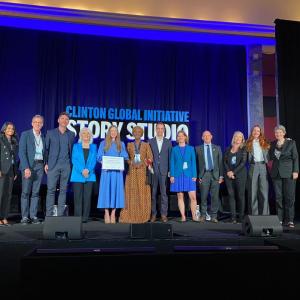 A row of people posed on a stage. One holding a plaque. "Clinton Global Initiative" projected behind them.