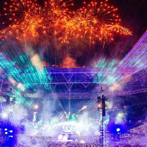 A huge stadium filled with spectators, a stage central is brightly lit, and orange fireworks fill the sky.