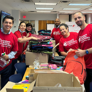 6 Aramark volunteers putting together backpacks, filling them up with assorted school supplies.