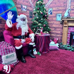 Santa waving at camera in front of Christmas tree and tablet