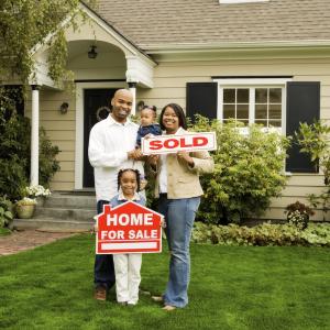 Young family of four in front of their new home holding a "sold" sign.