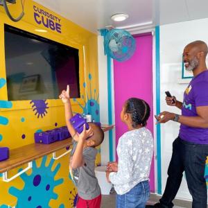 A smiling man stands next to two students who are engaging with interactive science materials. 