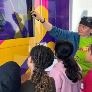 Volunteer inside the Curiosity Cube™ guiding a group of students through an interactive science activity on a TV screen.