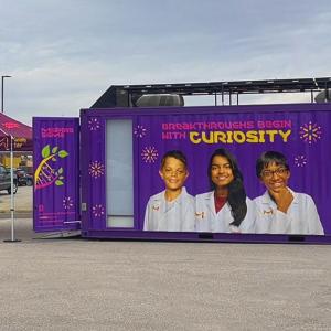 Exterior view of the Curiosity Cube™ mobile science lab featuring the message “Breakthroughs Begin with Curiosity” and an image of three student scientists.