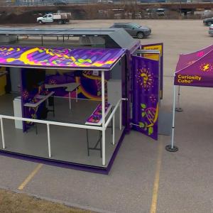 Aerial view of the Curiosity Cube™ mobile science lab with side panels open, revealing interactive experiment stations beneath a colorful canopy.