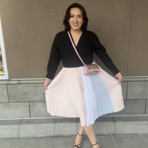 Curtlynn shows off her skirt, pleated pastel striped, strapy black heels and black v-neck top. She stands, smiling, in front of an exterior beige wall.