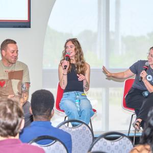 Three speakers sit onstage during a panel discussion, holding microphones and speaking to an audience seated in a conference room.
