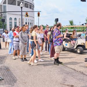 College program participants gather outdoors near crowd-control barricades as a staff member provides a venue operations briefing outside a stadium before an event.